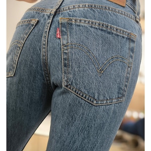Könnte beinhalten: Nahaufnahme einer blauen Jeans mit klassischem Fünf-Taschen-Design. Die Jeans hat ein rotes Etikett auf der Gesäßtasche und einen braunen Lederaufnäher. Die Nähte sind sichtbar, und die Jeans scheint abgenutzt zu sein.