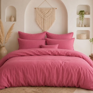 May include: A bed with a magenta-colored duvet cover and matching pillowcases. The bedding is arranged on a bed frame, with a neutral-toned rug beneath. The room has a bohemian aesthetic, with decorative elements like a macrame wall hanging and pampas grass in a vase.