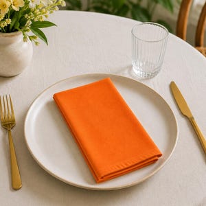 May include: A table setting with a white plate, orange napkin, gold fork and knife, and a clear glass. A small vase with yellow flowers sits to the left. The table is covered with a white tablecloth.