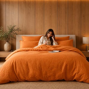 Pode incluir: Um conjunto de cama laranja com fronhas e uma capa de edredão a condizer. A roupa de cama está numa cama com cabeceira de madeira e uma mulher sentada na cama. Uma lâmpada e uma planta estão nas mesas de cabeceira.