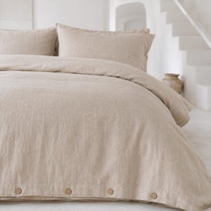 Puede incluir: Juego de cama de lino beige, que incluye una funda nórdica y dos fundas de almohada. La funda nórdica tiene botones de madera en el borde inferior. El juego está hecho de una tela texturizada, creando una estética relajada. La paleta de colores neutros complementa varios estilos de dormitorio.