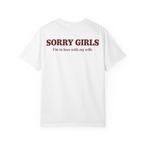 May include: A white short-sleeved t-shirt featuring dark red text printed on the back. The text reads SORRY GIRLS in a bold, capitalized font, followed by the smaller, centered text I'm in love with my wife in a simple serif typeface.