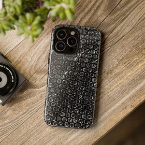 May include: Black phone case with a bubble wrap pattern. The case is on a wooden surface, with a camera and a plant in the background. The case has a glossy finish and a textured appearance.