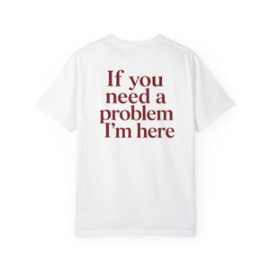 May include: A white short-sleeved t-shirt featuring dark red text printed on the back. The text reads, "If you need a problem I'm here" in a serif font. The garment has a crew neckline and a standard fit, showcasing the back view of the shirt.