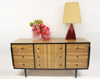 May include: A mid-century modern dresser with six drawers, black trim, and black legs. The dresser is made of wood with a light brown finish. A pink vase with pink flowers sits on top of the dresser, and a red lamp with a yellow lampshade sits on the right side.