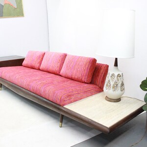 May include: A mid-century modern sofa with a pink and orange striped fabric upholstery. The sofa has a built-in side table with a drawer and a separate side table with a white marble top.