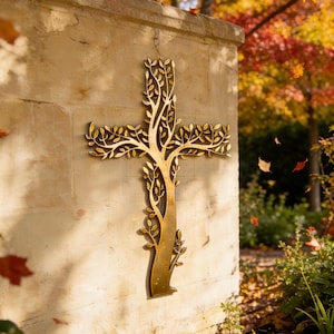 May include: A gold-colored metal cross with a tree-like design, mounted on a textured beige wall. The cross features branches and leaves, with a natural, organic aesthetic. The background includes autumn foliage.