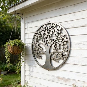 Peut inclure: Une décoration murale ronde en métal représentant un arbre de vie avec un design découpé détaillé. L'arbre présente un tronc, des branches et des feuilles dans un cadre circulaire. Il est monté sur un mur en bois blanc, avec un panier suspendu de plantes à proximité.