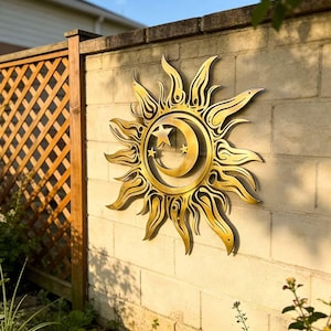 Peut inclure: Une œuvre d'art murale en métal doré représentant le soleil et la lune. Le soleil a des rayons détaillés, et la lune présente un design d'étoile et de croissant. L'œuvre est montée sur un mur de briques de couleur claire, avec une clôture en bois et de la verdure en arrière-plan.