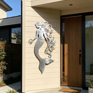 Puede incluir: Escultura de pared de metal plateado de una sirena. La sirena tiene cabello suelto y una cola detallada, montada en una pared exterior beige. Cerca hay una puerta de madera y una entrada de hormigón.