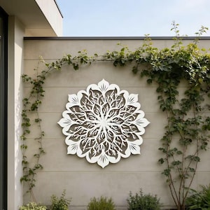 Puede incluir: Arte mural floral de metal blanco cortado con láser. La pieza decorativa está montada en una pared de color claro, con enredaderas y vegetación trepadoras. La obra de arte añade un toque moderno al espacio exterior.