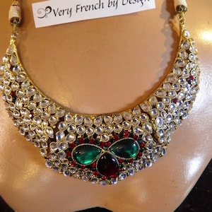 May include: A gold necklace with a crescent shape design. The necklace is adorned with clear, round stones and red and green gemstones. The necklace has a tag that reads "Very French by Design."