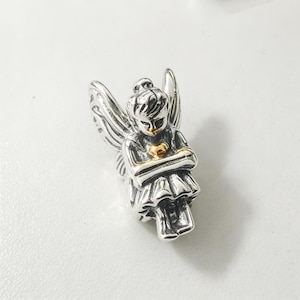 Puede incluir: Un dije de plata con forma de hada sentada con alas detalladas, que lleva un vestido y sostiene un pequeño corazón de color dorado contra el pecho. El metal tiene un acabado pulido con áreas oscurecidas para resaltar el diseño intrincado.