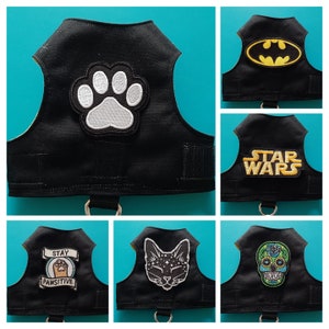 Könnte beinhalten: Sechs schwarze Hundegeschirre mit verschiedenen gestickten Aufnähern. Die Aufnäher beinhalten einen Pfotenabdruck, ein Batman-Logo, die Worte "Star Wars", ein Katzen Gesicht, einen Schädel mit Blumen und einen Pfotenabdruck mit den Worten "Stay Pawsitive".