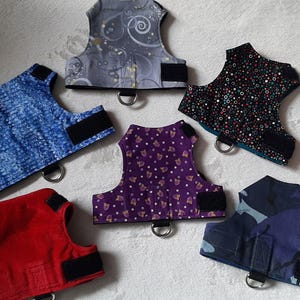 Op de afbeelding: Zes verschillende hondenharnassen in verschillende kleuren en patronen. De harnassen zijn gemaakt van stof en hebben verstelbare banden.