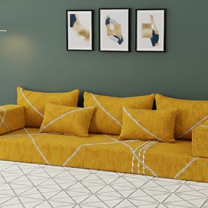 Puede incluir: Un sofá modular amarillo mostaza con costuras geométricas blancas. El sofá tiene varias almohadas a juego. El fondo presenta arte abstracto enmarcado y una lámpara de pie dorada. El sofá está sobre una alfombra estampada.