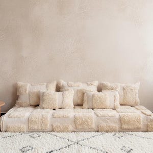 May include: A cream-colored, tufted floor cushion with matching square pillows. The cushion and pillows have a textured, raised pattern. A small, round wooden table is to the left, and a white rug with a geometric pattern is in the foreground.
