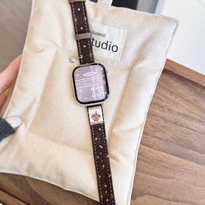 May include: A brown watch band with white star patterns and a small charm. The watch face displays the time 3:34 and the word "Starlight". The band is on a beige padded surface with the word "studio" printed on it.
