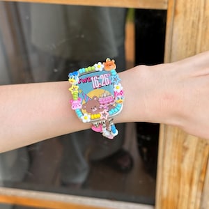 May include: A colorful wristwatch with a birthday theme. The watch face displays the time 16:26 and features a cartoon bear, cake, and other decorative elements. The watch is adorned with various charms and a light blue strap.