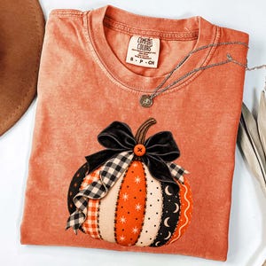 Puede incluir: Una camiseta naranja de manga corta con cuello redondo y etiqueta Comfort Colors. El gráfico muestra una calabaza hecha de paneles de tela verticales con patrones en naranja, negro y blanco, rematada con un lazo de terciopelo negro y botón.