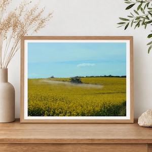 May include: A framed print featuring a vibrant field of yellow flowers under a blue sky. A green tractor is visible in the distance, spraying the field. The print is in a wooden frame, resting on a wooden surface.