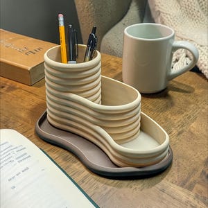 May include: A beige desk organizer with a wavy design, holding pens and a pencil. A matching mug sits nearby. The organizer is on a brown tray, with a book and open notebook in the background. The desk organizer is a stylish addition to any workspace.