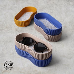 May include: A collection of four oval-shaped storage trays with ribbed exterior walls and grey felt-lined interiors. The trays are colored mustard yellow, blue, beige, and dark blue. A pair of black sunglasses rests inside the beige tray. Text reads Crooze Creations Limited.