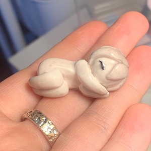 Puede incluir: Una pequeña figura de color blanquecino de un bebé durmiendo. La figura está acostada de lado con los ojos cerrados. El bebé lleva un pequeño gorro. La figura se sostiene en una mano.