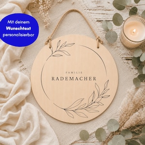 May include: A round light-colored wooden sign with a jute rope hanger. The sign features a black line-art botanical wreath design surrounding the centered text "FAMILIE RADEMACHER" in a clean, sans-serif font, with a blue circular overlay.