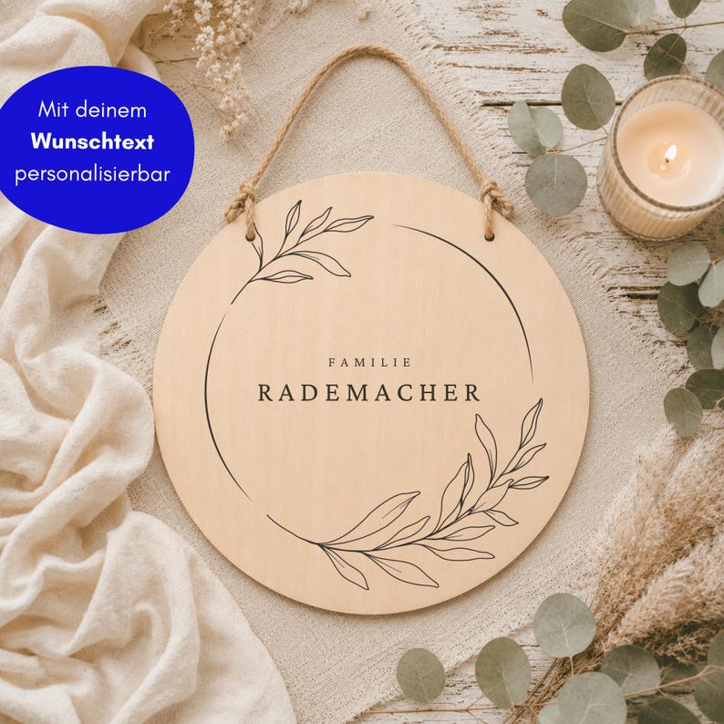 May include: A round light-colored wooden sign with a jute rope hanger. The sign features a black line-art botanical wreath design surrounding the centered text "FAMILIE RADEMACHER" in a clean, sans-serif font, with a blue circular overlay.