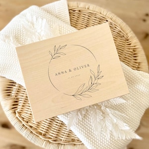 K&ouml;nnte beinhalten: Eine rechteckige, helle Holzschachtel mit einem schwarzen Strichzeichnungs-Design eines kreisf&ouml;rmigen Kranzes mit Bl&auml;ttern. Der Text ANNA & OLIVER ist mittig aufgedruckt, wobei das Datum 11.05.2024 direkt unter den Namen in schwarzer Tinte steht.