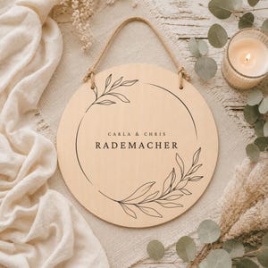May include: A round light-colored wooden sign featuring a black line-art botanical wreath design. The text "CARLA & CHRIS" is printed above the name "RADEMACHER" in the center. The sign includes a jute rope hanger attached to the top.