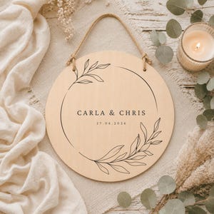 May include: A circular light-colored wooden sign featuring a black line-art wreath design. The text "CARLA & CHRIS" is printed in the center, followed by the date "27.06.2026". The sign includes a jute rope hanger attached to the top for display.