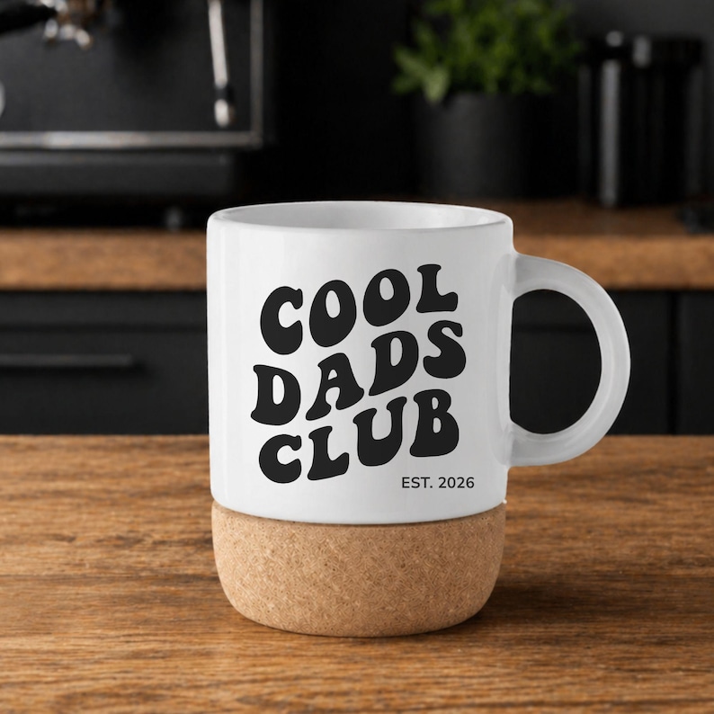 May include: White ceramic mug with the text "COOL DADS CLUB" in black, with "EST. 2026" below. The mug sits on a cork base, and has a curved handle. The mug is on a wooden surface.