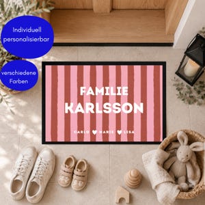 Könnte beinhalten: Eine rechteckige Fußmatte mit schwarzem Rand und einem Muster aus vertikalen rosa und terrakottafarbenen Streifen. Der Text FAMILIE KARLSSON ist mittig in Weiß gedruckt, darunter die Namen CARLO, MARIE und LISA, getrennt durch weiße Herzsymbole.