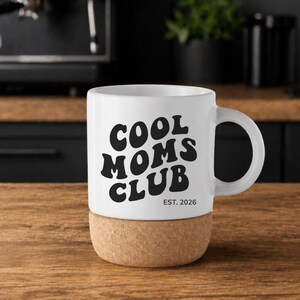 May include: White ceramic mug with the text "COOL MOMS CLUB" in black, with "EST. 2026" below. The mug sits on a cork base and has a curved handle. The mug is on a wooden surface.