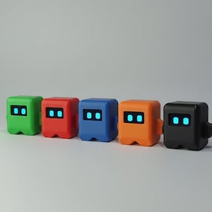 Può includere: Cinque giocattoli robot colorati in verde, rosso, blu, arancione e nero. Ogni robot ha una testa quadrata con due occhi blu luminosi. I robot sono a forma di cubo con bordi arrotondati e piccole sporgenze laterali.