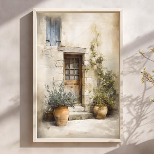 May include: A framed watercolour print depicting a European building with a wooden door and blue shutters. Two terracotta pots with plants sit either side of the entrance. The artwork is framed in a light-coloured wooden frame, creating a sense of calm.