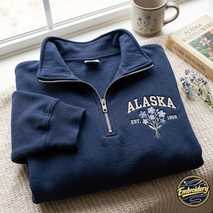 May include: A navy blue quarter-zip sweatshirt featuring an embroidered design on the chest. The embroidery includes the text ALASKA in an arched font, the text EST. 1959, and a small bouquet of light blue forget-me-not flowers with green stems.