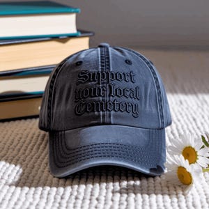 May include: A charcoal grey baseball cap with a distressed finish, featuring the embroidered black text Support your local Cemetery in a gothic font style. The cap has a structured front panel, a curved brim, and visible stitching details.