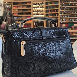 Puede incluir: Un bolso de mano negro con un patrón texturizado de piel de serpiente, que cuenta con un asa superior, un tirador de cremallera dorado en el bolsillo trasero y herrajes dorados para una correa de hombro en los laterales, de forma rectangular.
