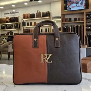 May include: A rectangular leather briefcase featuring a two-tone design with a smooth tan left side and a textured dark brown right side. The bag includes two top handles and a prominent gold-toned metal logo with the letters RZ centered on the front.