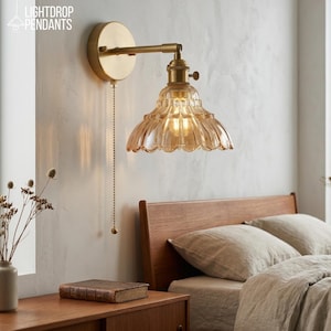 Könnte beinhalten: Eine Wandleuchte mit rundem Messingsockel, verstellbarem Arm und einem gewellten, bernsteinfarbenen Glasschirm. Eine Metallzugkette hängt vom Sockel herab. Der Text LIGHTDROP PENDANTS ist in der oberen linken Ecke des Bildes zu sehen.