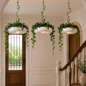 Op de afbeelding: Drie witte, komvormige hanglampen met groene klimop. De lampen hangen aan het plafond en verlichten de ruimte eronder. De neerhangende klimop voegt een natuurlijk element toe aan het interieur.