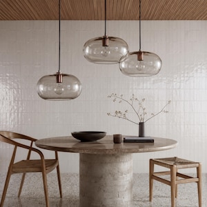 May include: Three pendant lights with rounded, transparent glass shades and copper-toned hardware hang above a circular stone dining table. The table is accompanied by a wooden chair with a woven seat and a small wooden stool, featuring a vase.