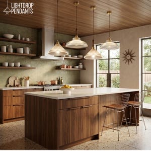 May include: A kitchen with wood cabinets, a white countertop, and three pendant lights. The lights have a clear glass shade and a brass finish. The kitchen also features a stove, shelves, and a kitchen island with two bar stools. The text "LIGHTDROP PENDANTS" is in the top left corner.