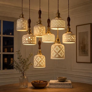 Op de afbeelding: Een hanglamp met zeven witte keramische kappen voorzien van diverse uitgesneden patronen. Elke kap hangt aan een bruin snoer met messing details en een houten accent, waardoor een warme gloed vanuit de lampen wordt verspreid.
