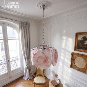 Könnte beinhalten: Eine moderne Hängeleuchte mit Chrom-Finish und einzigartigem Design. Die Leuchte hat einen zentralen Globus und umliegende rosa und weiße Glasscheiben. Sie wird an einer Kette aufgehängt und an der Decke befestigt. Der Text "LIGHTDROP PENDANTS" ist links oben sichtbar.