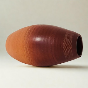 May include: An oval-shaped ceramic vase with a colour gradient. The vase transitions from light orange to deep burgundy. Textured bands encircle the vase. The opening is a dark, circular shape. The vase is approximately 30 cm long.