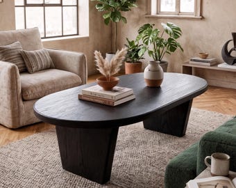 May include: A large, oval, black wooden coffee table with a thick top and two wide, angled pedestal legs. The table is positioned in a living room setting, featuring a beige armchair, a green sofa, and various decorative plants and books.